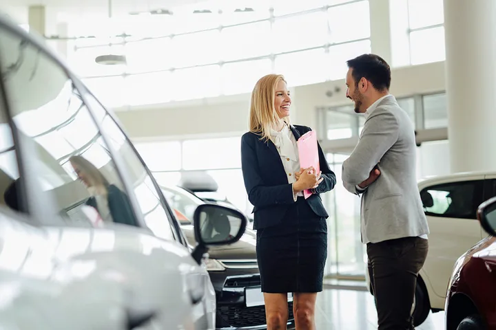 Female salesperson laughing and chatting with a male customer amid cars in a bright dealership.