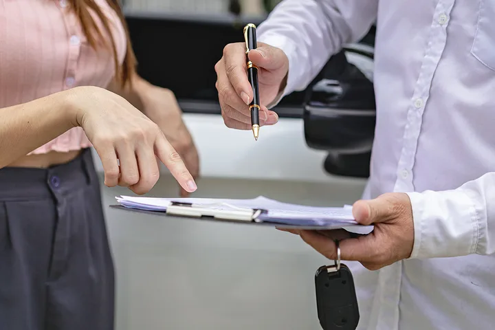 Salesperson holding car keys and clipboard while customer points at paperwork during a car deal.