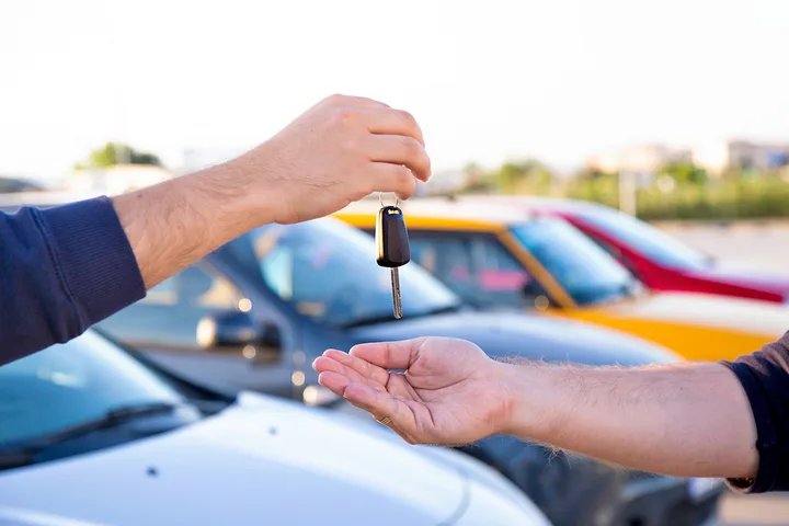 Two hands exchanging car keys in a parking lot with colorful cars in the background.