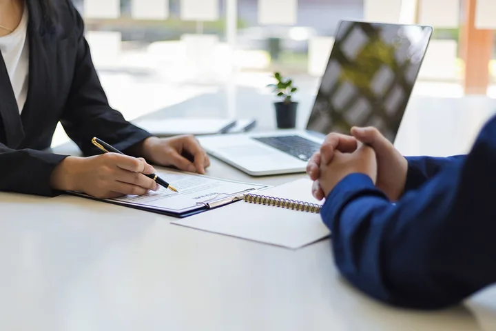 Two people at a desk reviewing paperwork with a laptop, in a formal meeting setting.