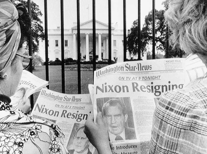 Black-and-white photo of two women reading 'Nixon Resigning' newspaper headlines outside the White House fence.