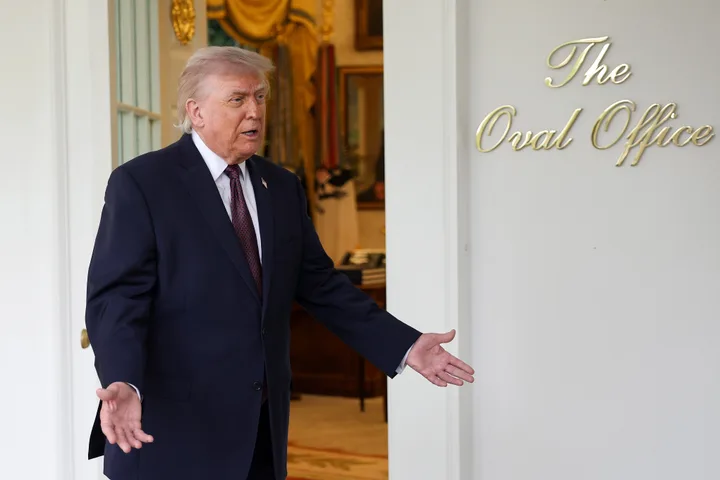 Trump standing in the doorway of the Oval Office gesturing outward, in a suit.