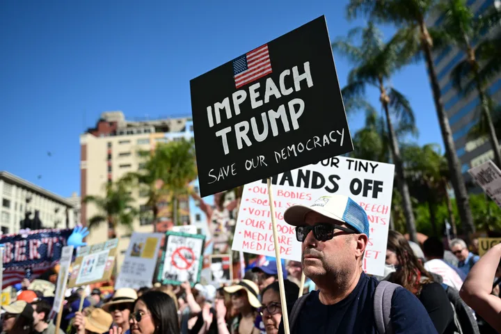 Large outdoor protest crowd with a prominent 'Impeach Trump Save Our Democracy' sign under blue sky.
