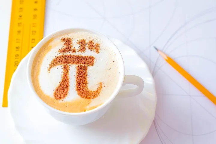 A latte in a white cup with a cinnamon pi symbol drawn in the foam, beside a ruler and pencil.