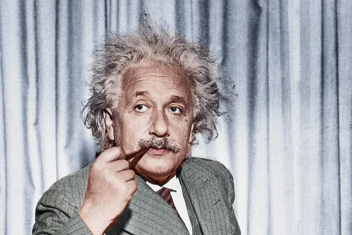 A colorized portrait of Albert Einstein looking thoughtful, holding a pen, against a grey curtain backdrop.