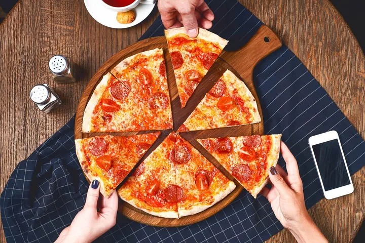 Hands reaching for slices of a round pepperoni pizza on a wooden board atop a restaurant table.