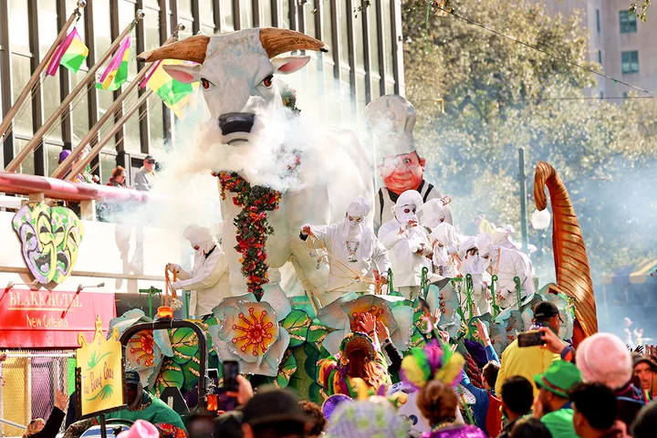 A large ox-head parade float with white-costumed riders emitting smoke rolls through a New Orleans street.