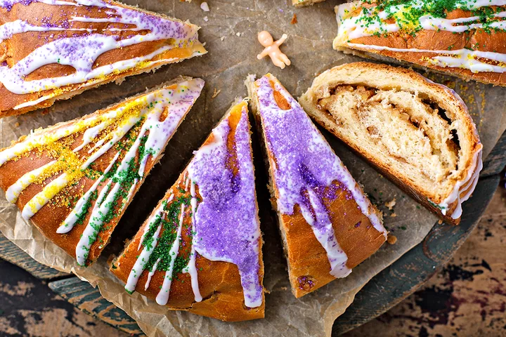 Sliced Mardi Gras king cake decorated with purple, green, and gold sugar and white icing on parchment.