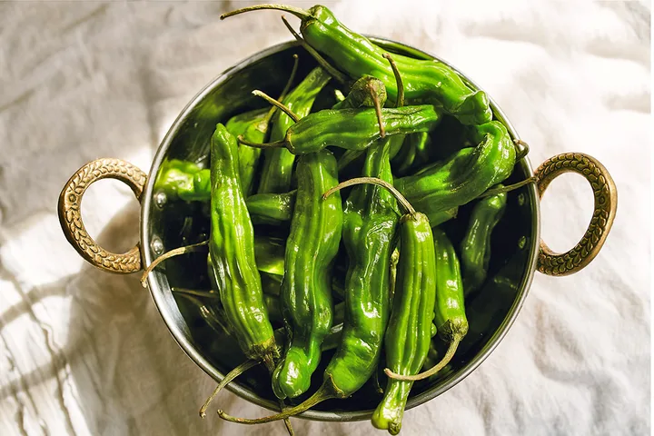 Fresh green shishito peppers piled in a decorative brass-handled metal bowl on white linen.