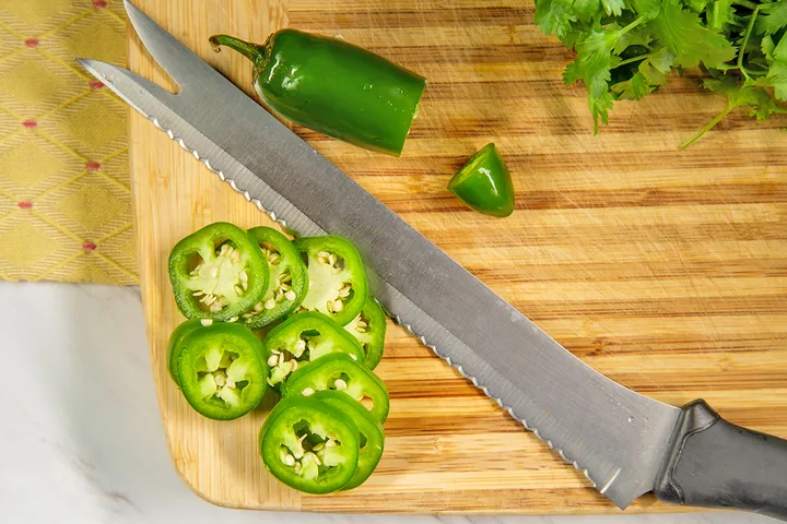 Sliced jalapeño pepper rounds and a whole jalapeño on a wooden cutting board with a serrated knife.