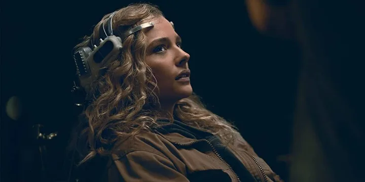 Woman with curly hair wearing a futuristic tech headset in a dark, moody sci-fi scene.
