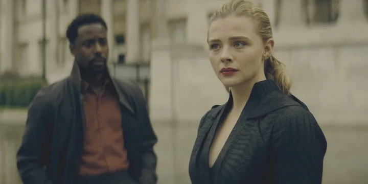 Two actors in a tense outdoor scene — a woman in black and a man in a leather jacket — with a brutalist building behind 