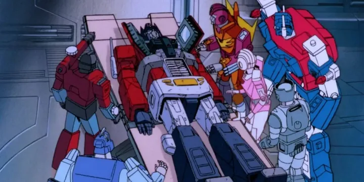 Multiple Transformers robots gather around a fallen comrade lying on a surface in an animated scene.