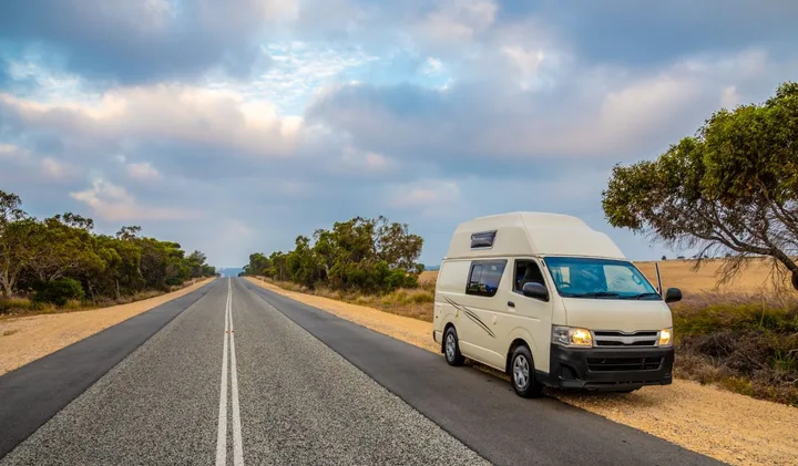 A white campervan driving along a straight empty road through flat Australian outback landscape.