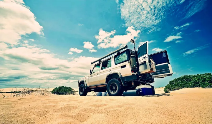 A rugged 4WD Land Cruiser parked on sand dunes under a dramatic blue sky, stylized photo.