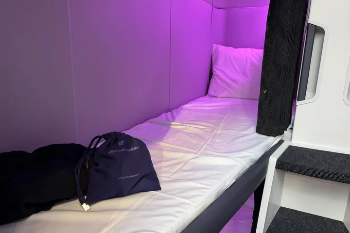Single airplane sleep pod with white pillow and bedding, purple lighting, and an Air New Zealand amenity bag.