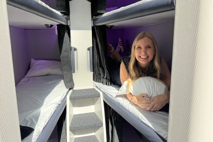 Smiling woman lying in an airplane bunk bed pod, hugging a pillow, with purple-lit sleeping berths visible.
