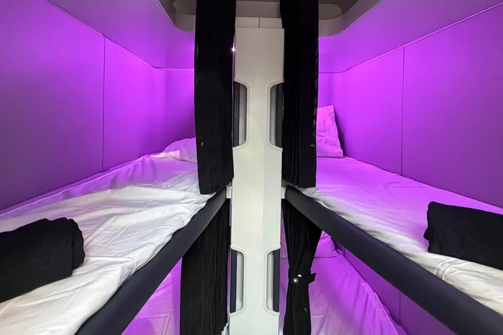 Interior view of stacked airplane sleep pods with white bedding, black curtains, and purple mood lighting.