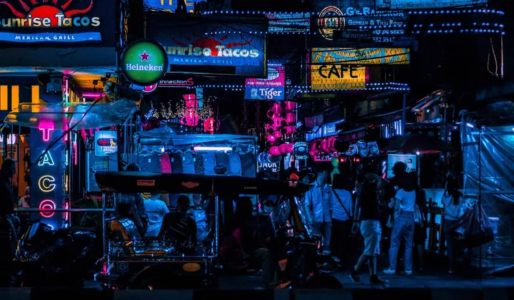 Neon-lit nightlife street in Bangkok with bars, signs, and crowds of people walking at night.