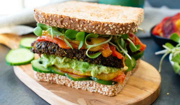 Stacked veggie sandwich with avocado, roasted peppers, and sprouts on seeded bread.
