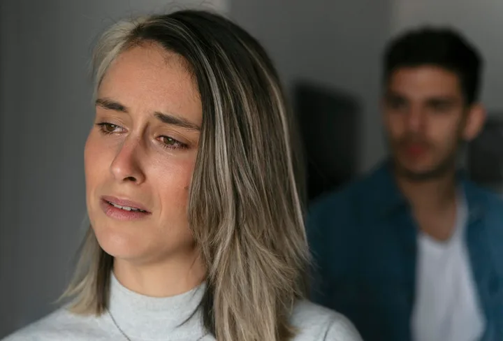 Distressed young woman with tears in eyes, man blurred in background, indoors.