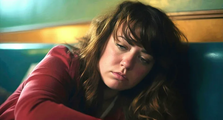 Sad young woman with dark hair sitting alone in a diner booth, looking downcast.