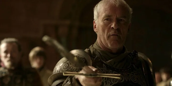 Older armored man holding an ornate weapon in a dimly lit hall with blurred crowd behind.