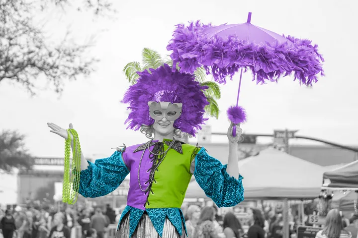 A woman in purple feathered Mardi Gras costume with mask and parasol holds beads at an outdoor festival.