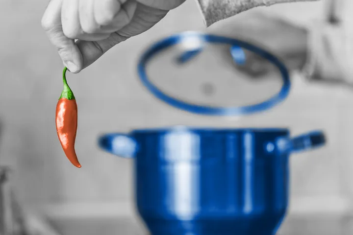 Hand holds a single red chili pepper over a blue cooking pot in a stylized selective-color kitchen photo.