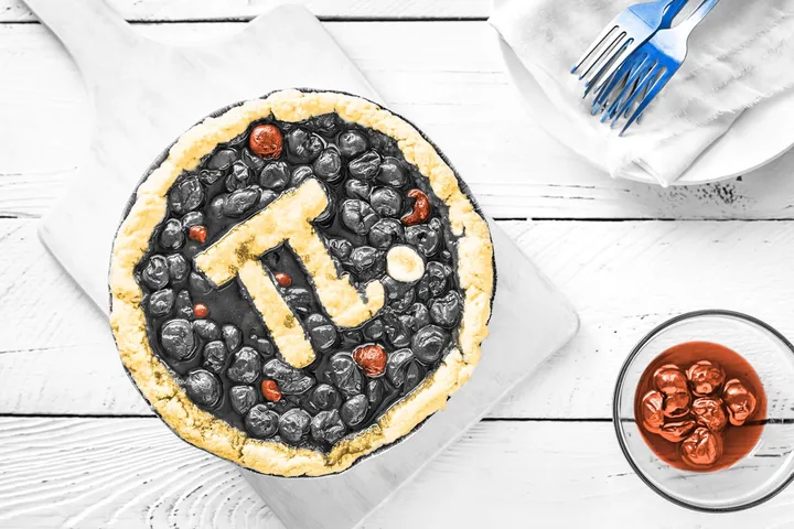 A dark berry pie with a golden pi symbol baked into the filling, shot from above on a white surface.