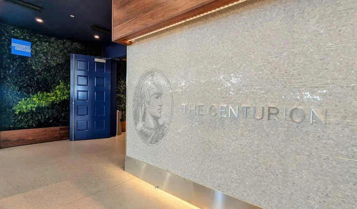 Entrance to the American Express Centurion airport lounge with mosaic logo wall and lush greenery.