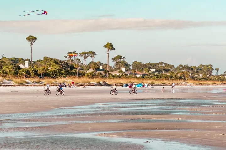 People riding bicycles and flying a kite on a wide sandy beach at golden hour.