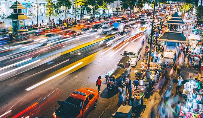 Aerial long-exposure shot of busy Bangkok street at night with light trails from traffic and street market.
