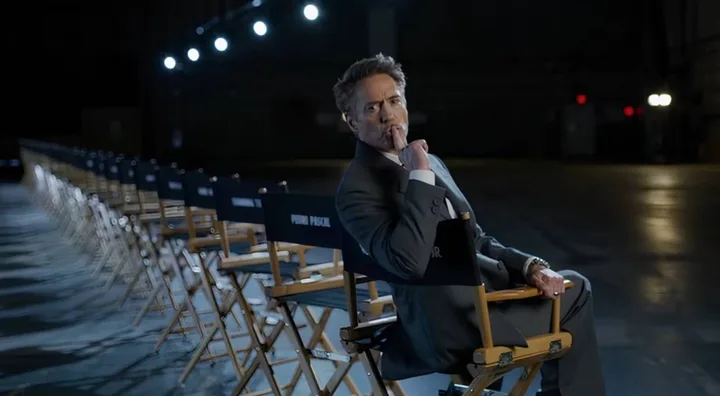 Man in suit sits pensively in a director's chair on a dimly lit film set with rows of empty chairs.