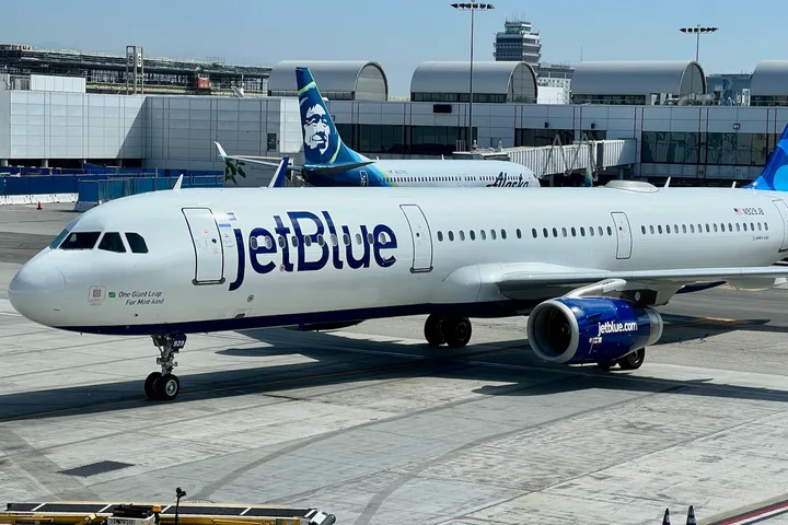 JetBlue aircraft taxiing on airport tarmac with Alaska Airlines plane visible in background.