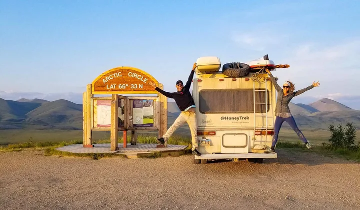 Two people jumping joyfully beside an RV at the Arctic Circle sign amid open mountain landscape.