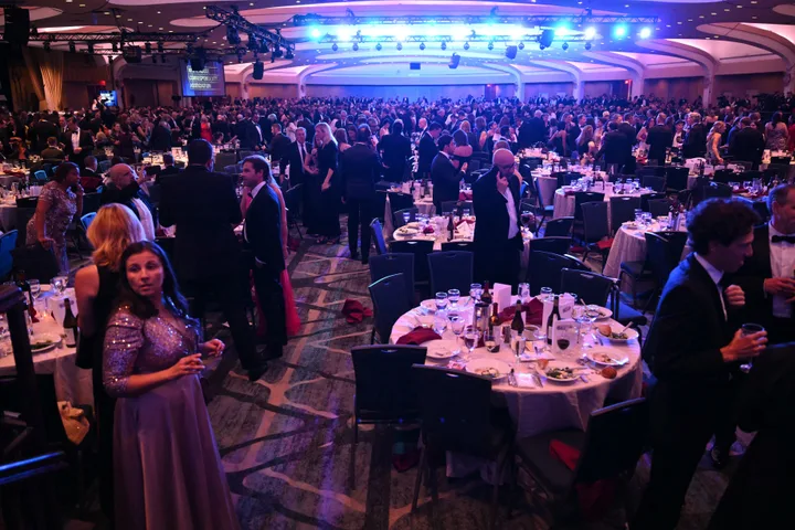 Large ballroom packed with formally dressed guests at a gala dinner with blue stage lighting.