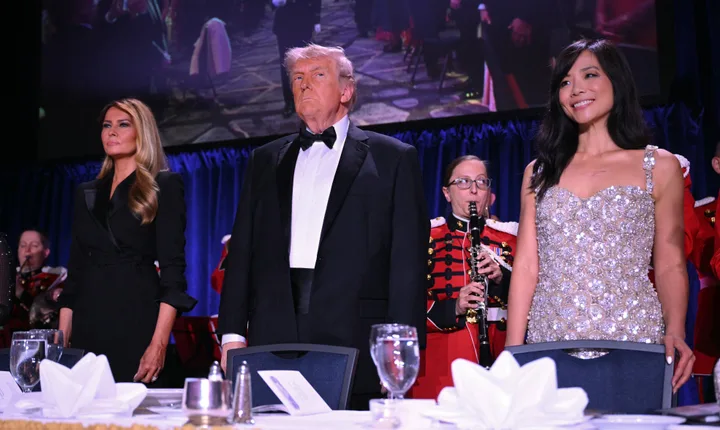 Donald Trump in tuxedo stands with two women at a formal dinner event with military band behind them.