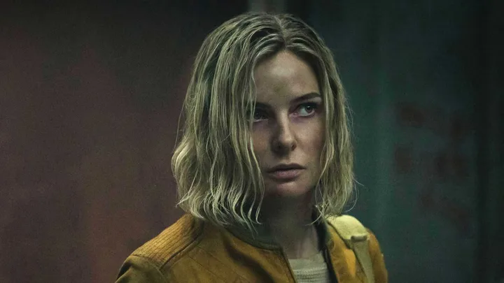 Woman with disheveled blonde hair in a yellow jacket looking tense in a dark, gritty cinematic setting.