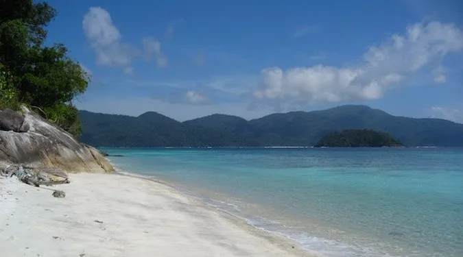 Pristine white sand beach with clear turquoise water and forested hills in background.
