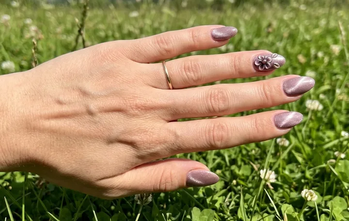 Close-up of a hand with almond-shaped mauve shimmer nails featuring a 3D flower accent, over green grass.