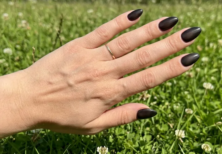 Close-up of a hand with long almond-shaped dark matte black nails, held over green grass.