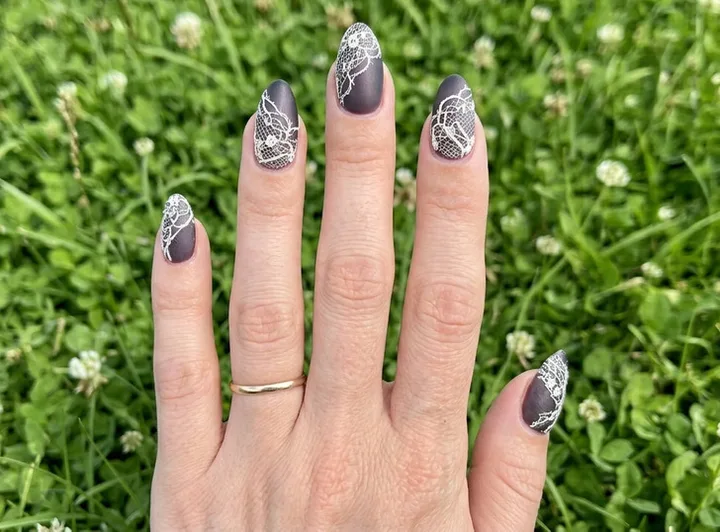 Close-up of a hand with almond nails featuring dark gray matte base and intricate white lace nail art over green grass.