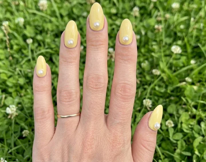 Close-up of a hand with almond-shaped yellow nails each adorned with a single white pearl, over green grass.