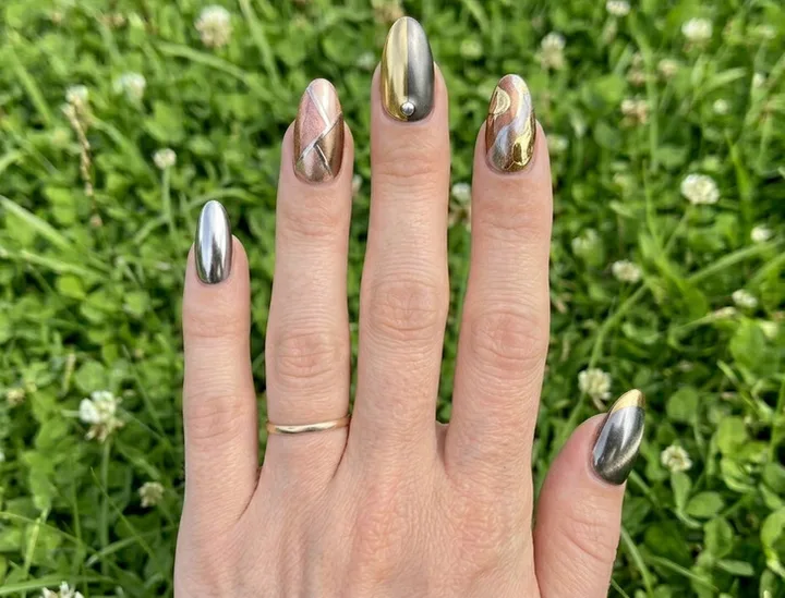 Close-up of a hand with mixed metallic chrome nails in silver, gold, and rose gold with abstract designs, over green gra