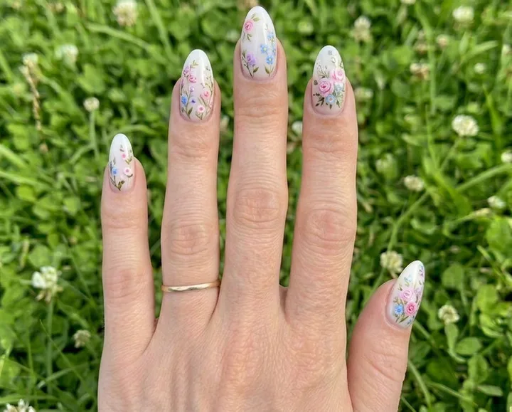 Close-up of a hand with almond nails painted with delicate pastel floral art on white base, over green grass.