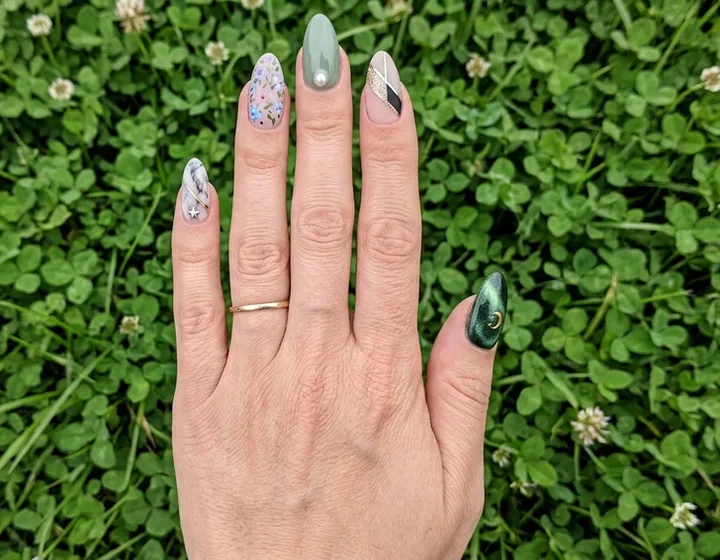 Close-up of a hand with mismatched nail art — sage green, floral, geometric, and dark chrome — over green clover.
