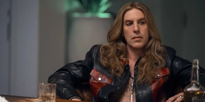 A man with long wavy hair in a red-and-black leather jacket seated at a table in a dimly lit scene.