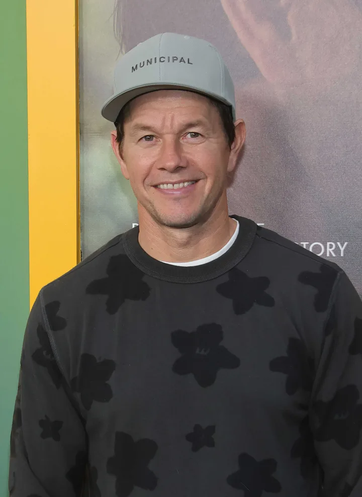 A smiling man in a cap and dark patterned sweatshirt posing at what appears to be a film premiere.