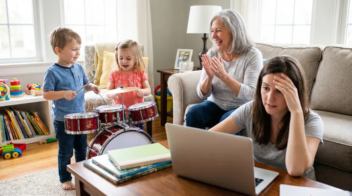 Frustrated woman at laptop while grandmother claps as two young children play a drum set in a living room.